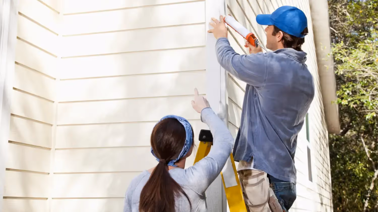 Two workers seal beige siding: one on a ladder applies caulk with a gun, while the other on the ground points to the seam.