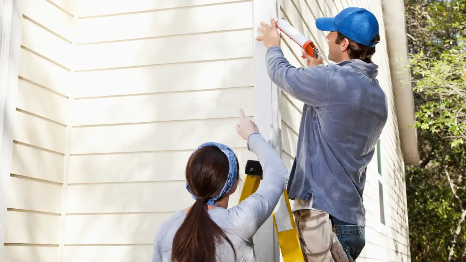 Two workers seal beige siding: one on a ladder applies caulk with a gun, while the other on the ground points to the seam.