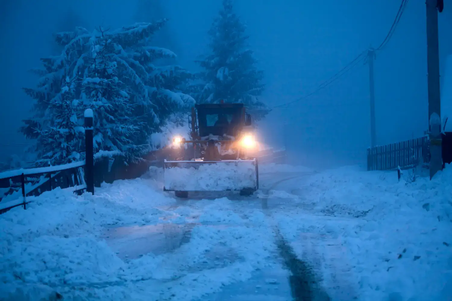 Snowplow clearing heavy snow on a rural icy road at dusk, with snow-covered trees, fence, and power lines in blue twilight