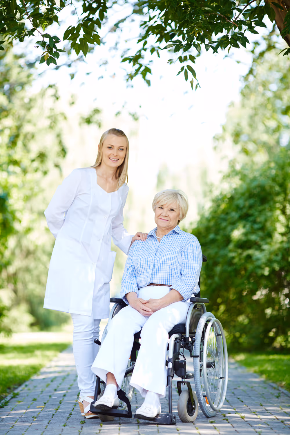 Young female caregiver standing outdoors behind a smiling elderly woman in a wheelchair on a paved path surrounded by greenery.