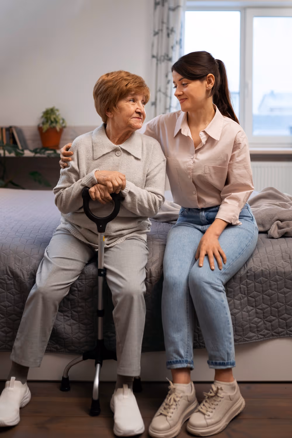 An elderly woman holding a walking cane sits on the edge of a bed, embraced and smiling warmly at a younger woman beside her.