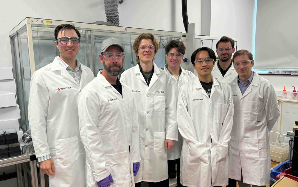 ReactWise scientists in white lab coats and safety glasses posing together inside a laboratory.