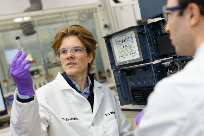 ReactWise Scientists in a lab coat and safety glasses holding a test tube in a laboratory setting.