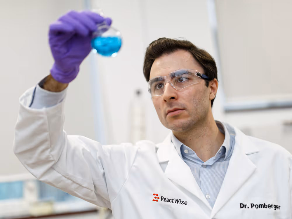 ReactWise Scientist wearing safety glasses and purple gloves holding a round-bottom flask with blue liquid in a laboratory.