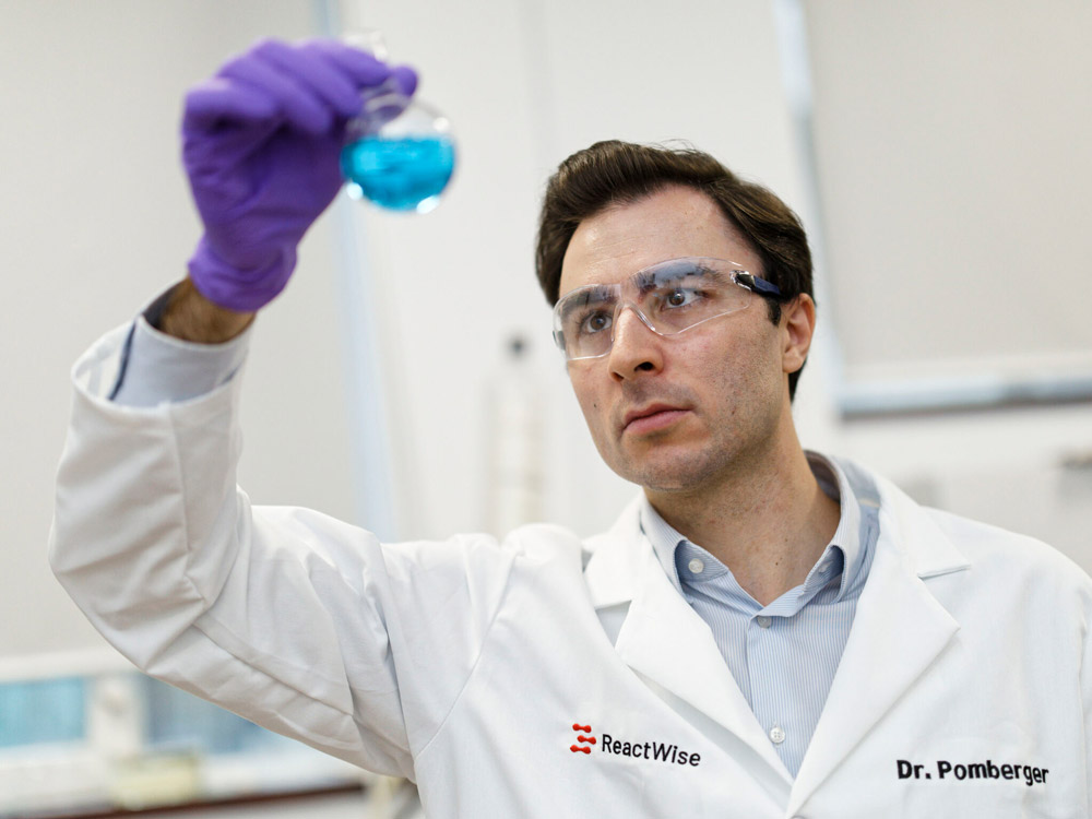 ReactWise Scientist wearing safety glasses and purple gloves holding a round-bottom flask with blue liquid in a laboratory.