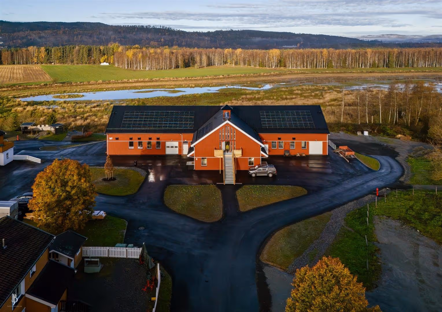 Rødt bygg med solcellepaneler på taket midt i et landlig område med høstfargede trær og åpne marker.