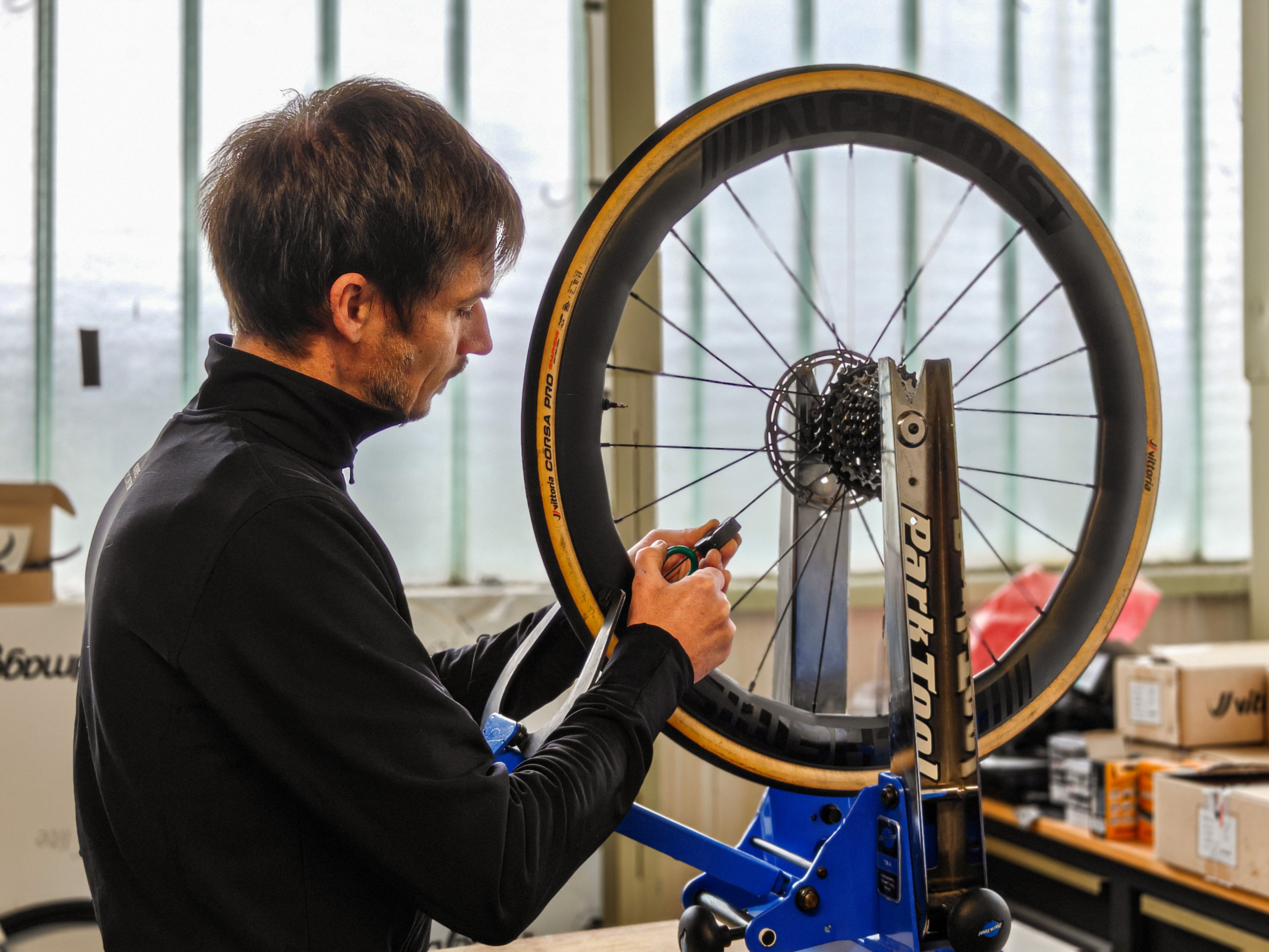 Man adjusting a bicycle wheel mounted on a blue Park Tool truing stand in a workshop.