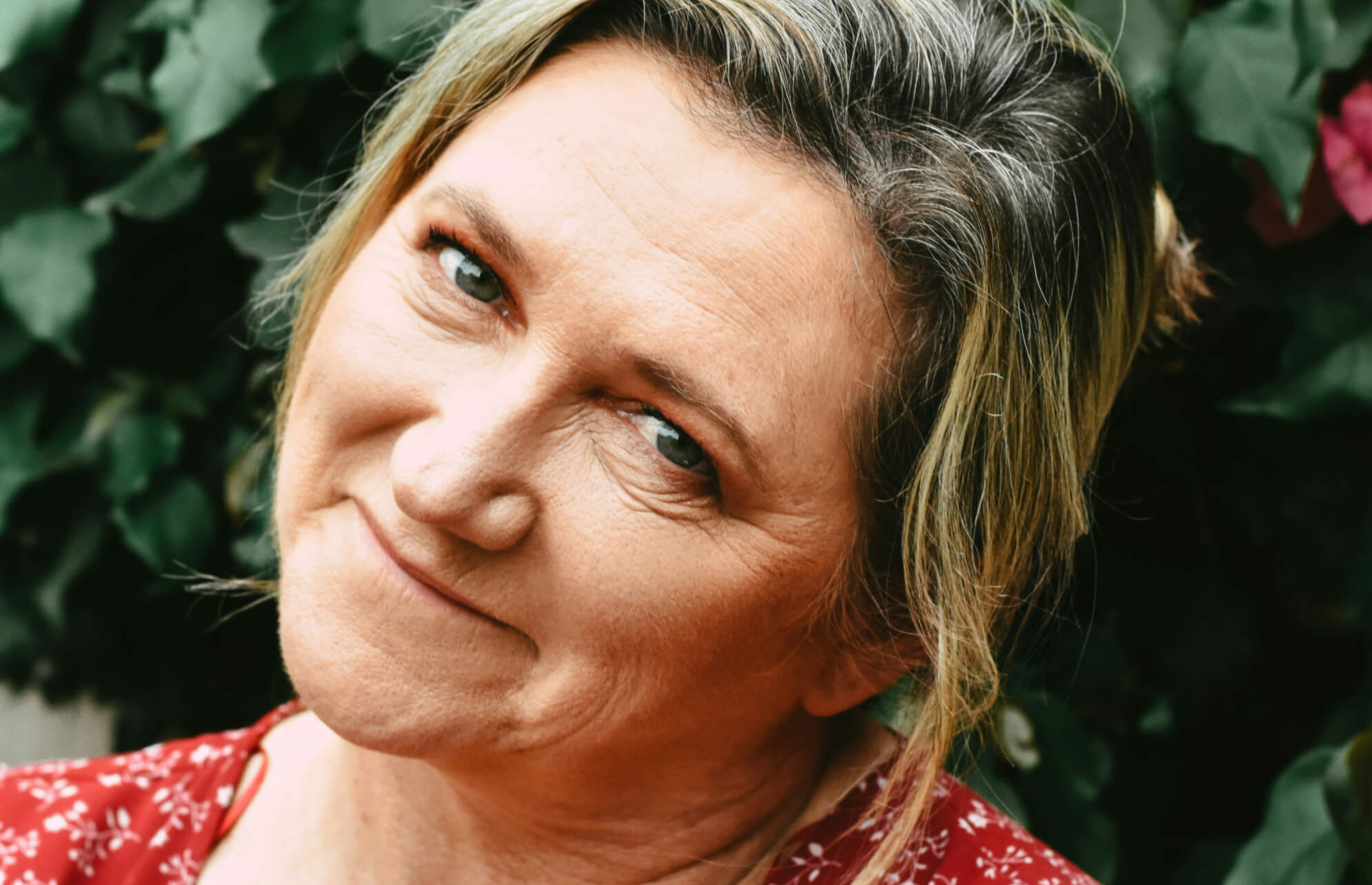 Close-up of a middle-aged woman with light hair smiling gently, wearing a red floral top, with a leafy background.