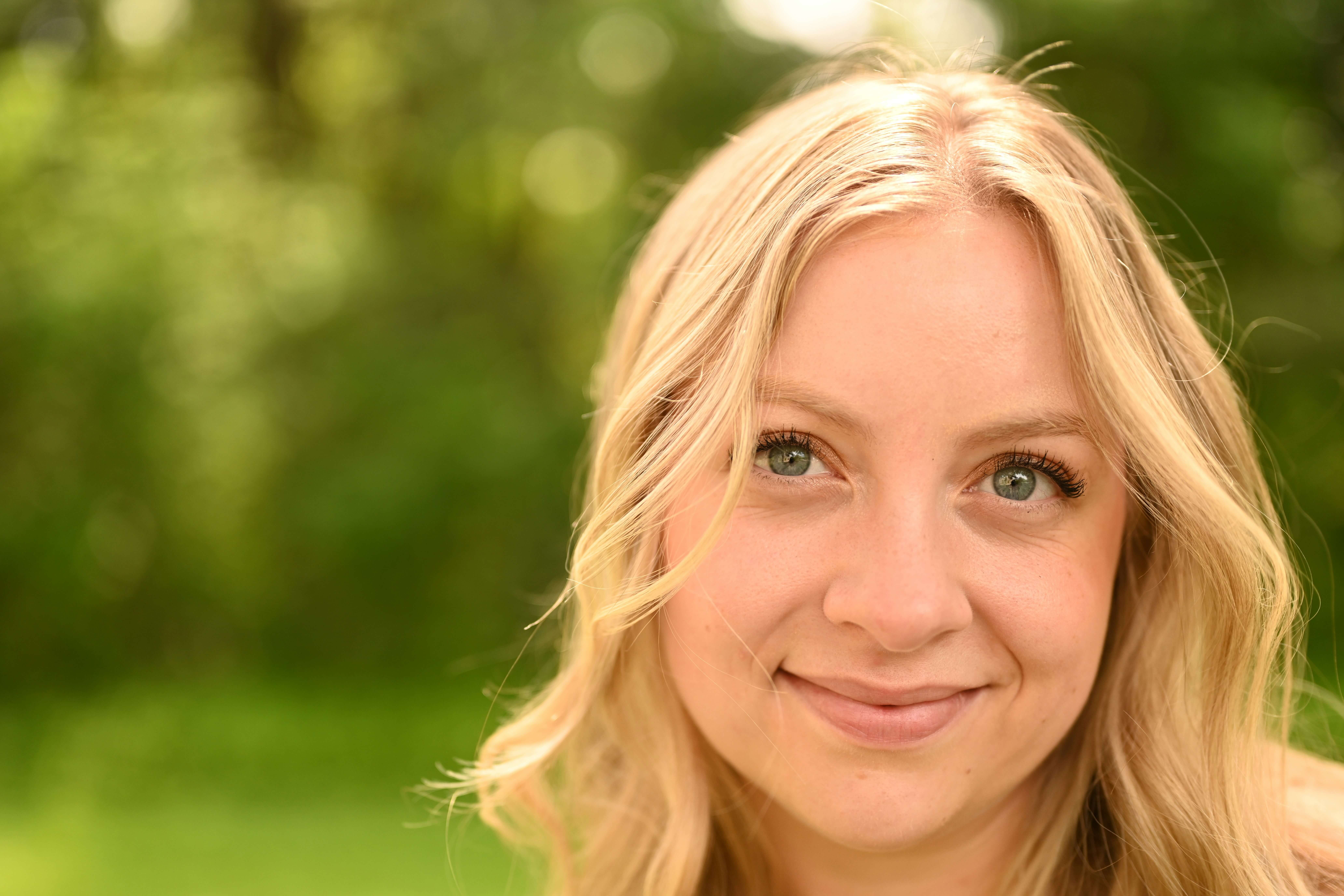 Close-up of a smiling young woman with blonde hair and green eyes outdoors with blurred green background.