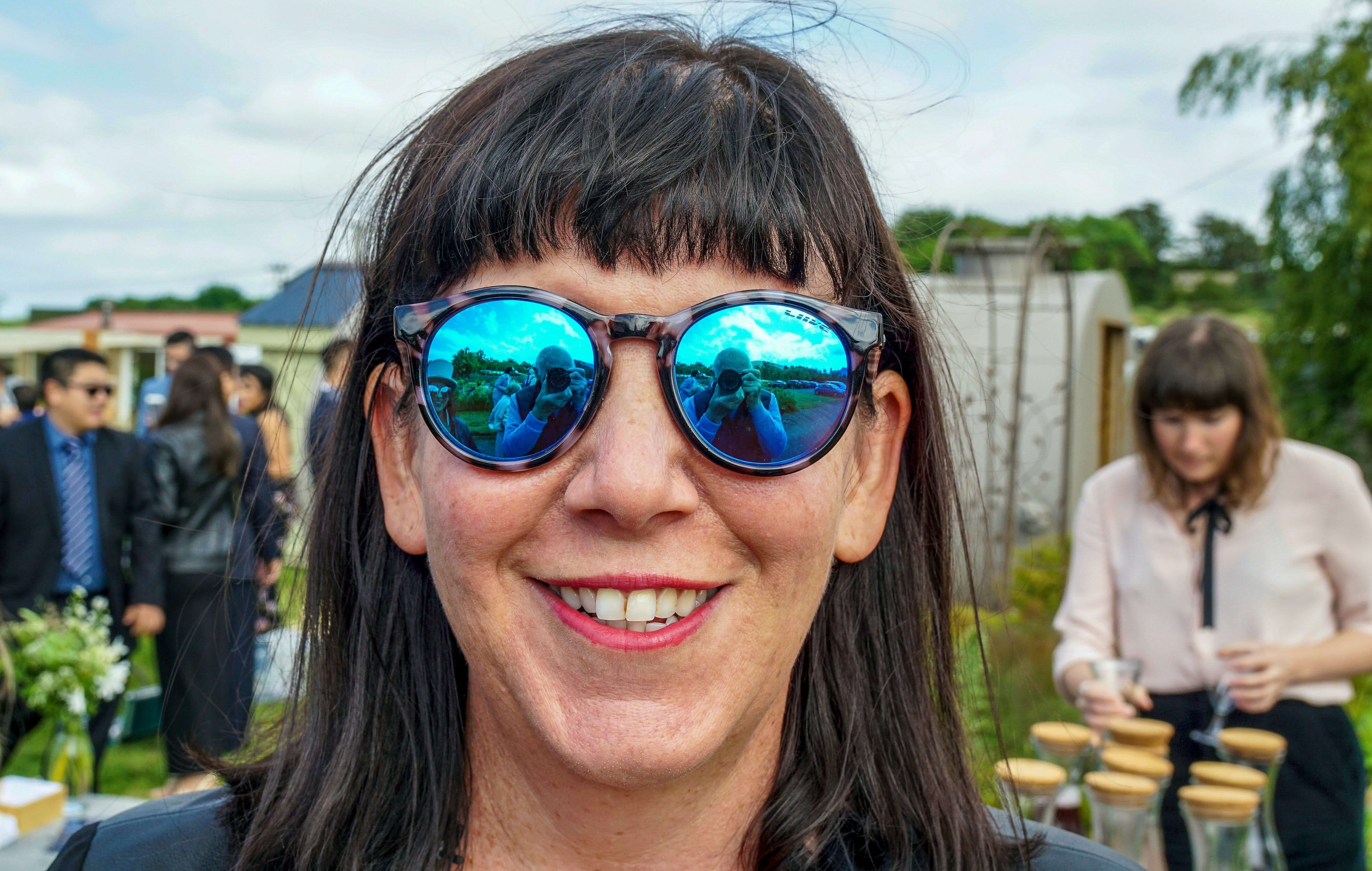 Close-up of a smiling woman wearing reflective blue sunglasses, with people and greenery in the background.