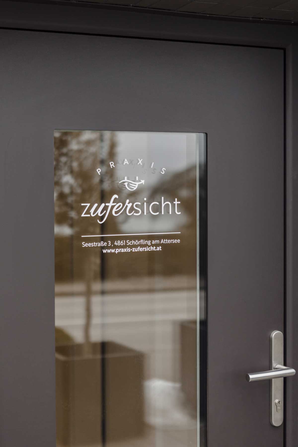 Eingangstür Praxis Zufersicht mit Spiegelung des Atterseeufers im Glas.
