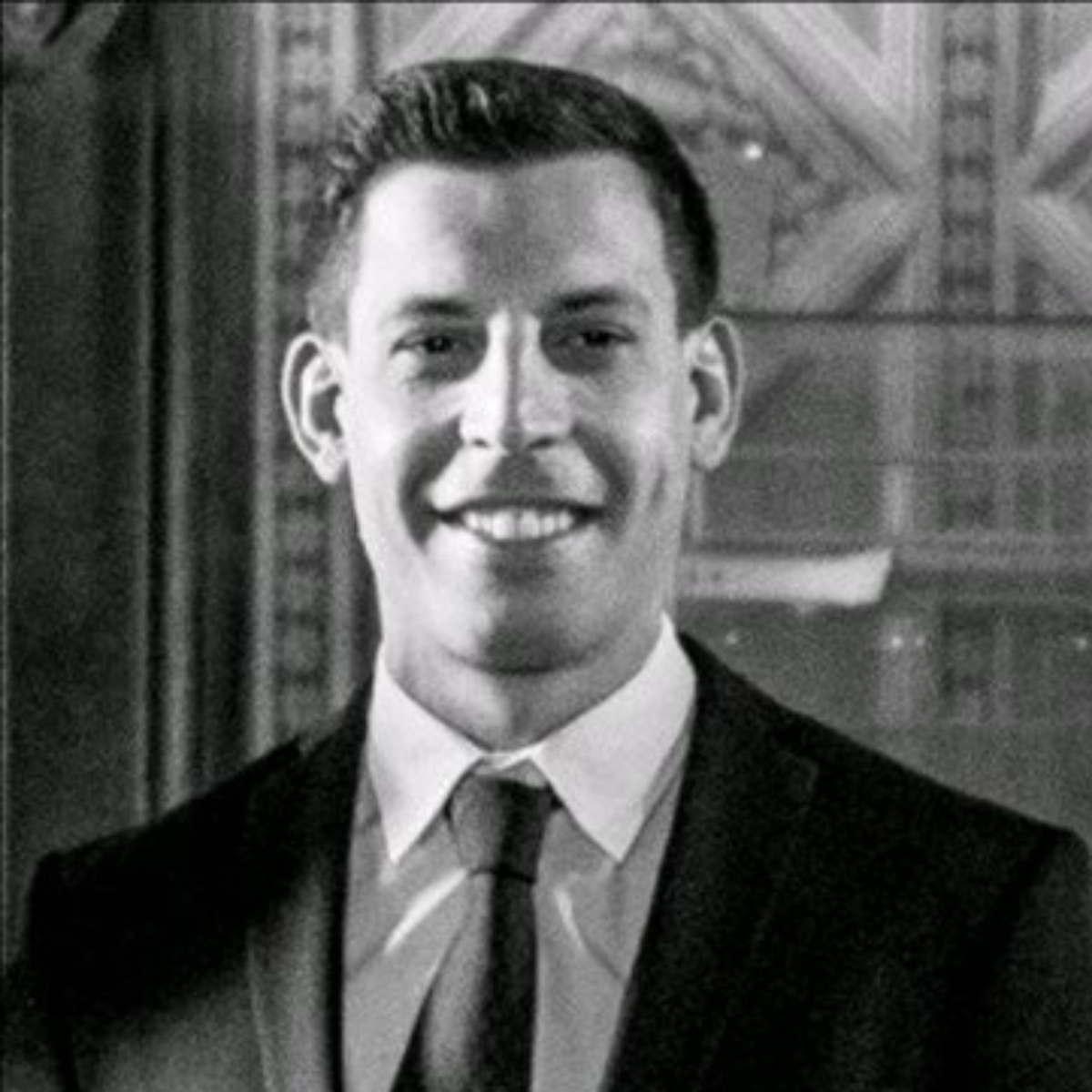 Black and white portrait of a smiling man i(Owner of the practice) n a suit and tie with a patterned background.