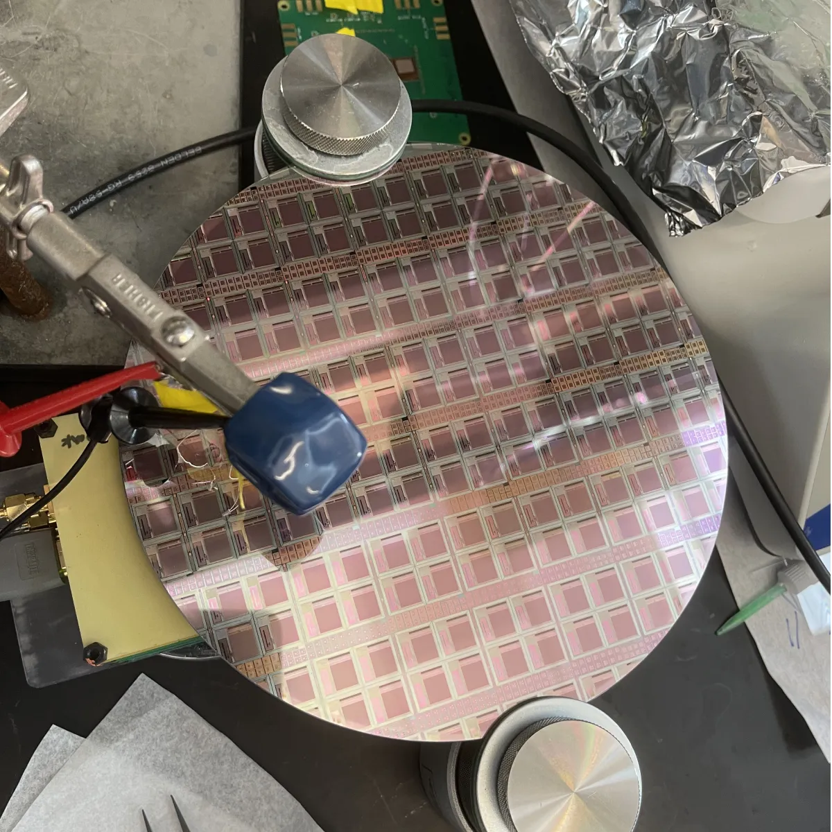 Close-up of a silicon wafer with rectangular microchips arranged in a grid pattern, held by a clip and surrounded by metal and electronic equipment.