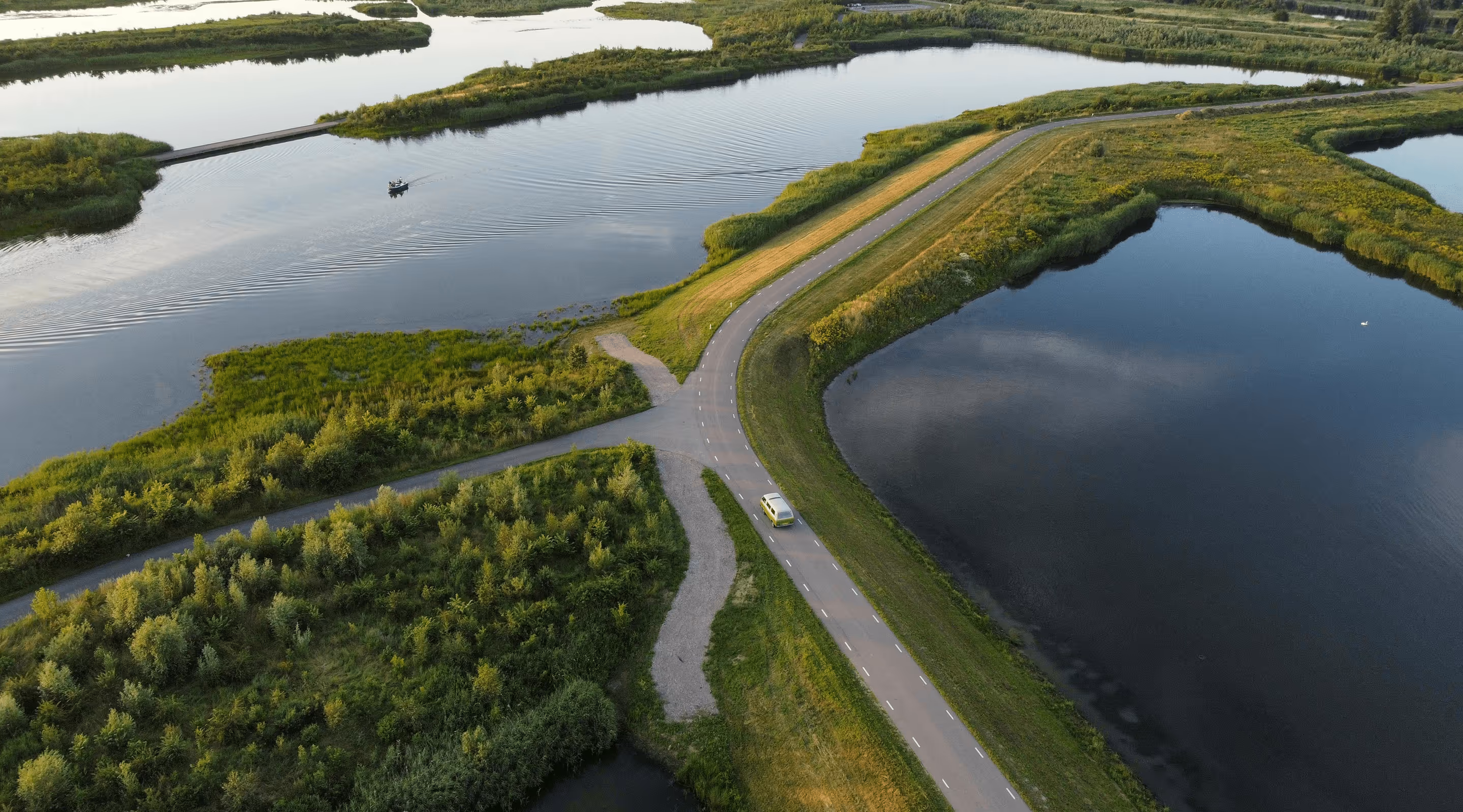 Luchtfoto van een kronkelende verharde weg omgeven door groene vegetatie en grote waterlichamen, met een klein geel voertuig op de weg en een boot op het water.