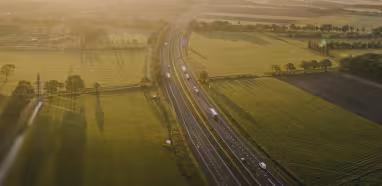 Luchtfoto van een snelweg die door groene en bruine velden loopt onder een felle zon tijdens de vroege ochtend of late namiddag.