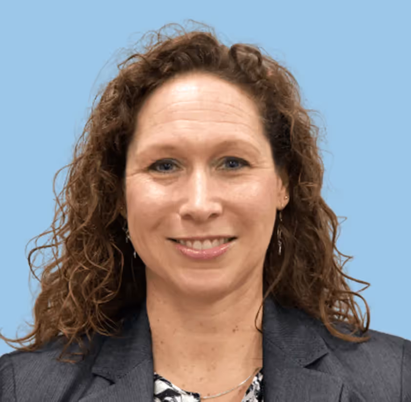 Smiling woman with curly brown hair wearing a gray blazer and patterned blouse against a light blue background.
