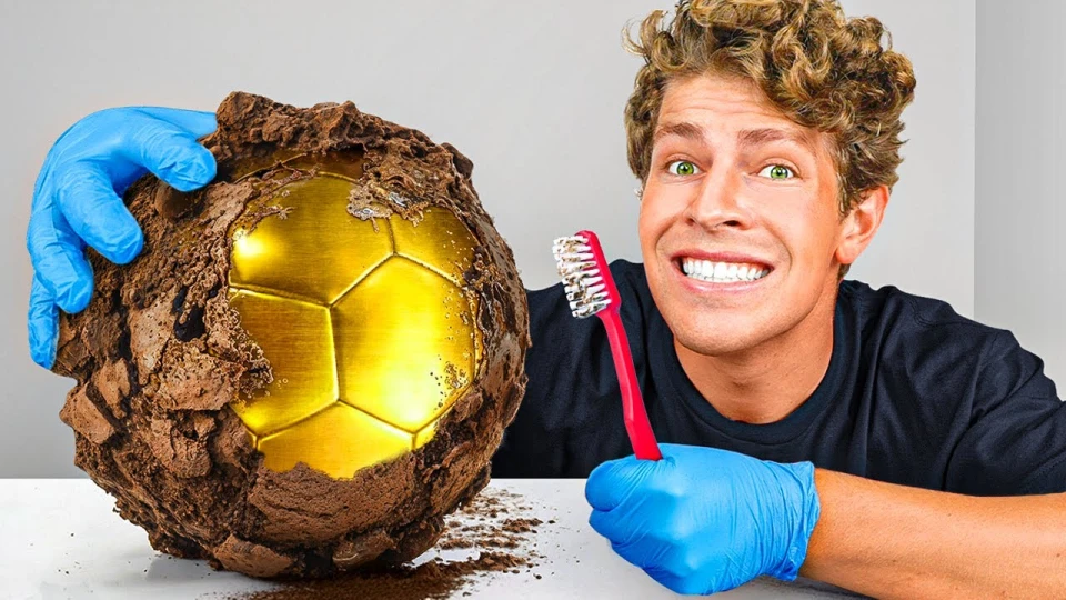 Man with curly hair wearing blue gloves smiles broadly, holding a red toothbrush beside a partially unearthed golden soccer ball covered in dirt on a white surface.