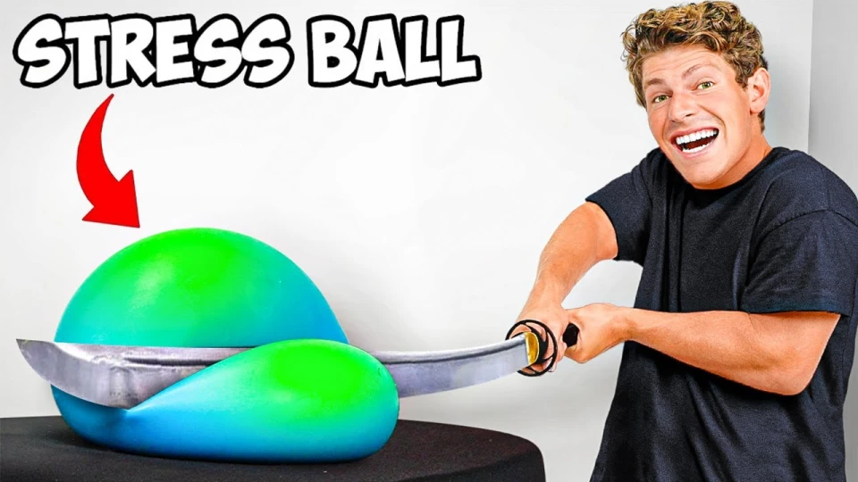 Man with curly hair in a black shirt smiling widely as he presses down on a large green and blue stress ball with a curved dagger, causing the stress ball to deform on a black surface.