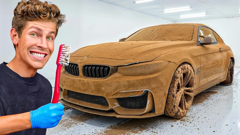 Man wearing a black shirt and blue glove smiling while holding a large red toothbrush next to a luxury car covered entirely in thick mud inside a white garage.