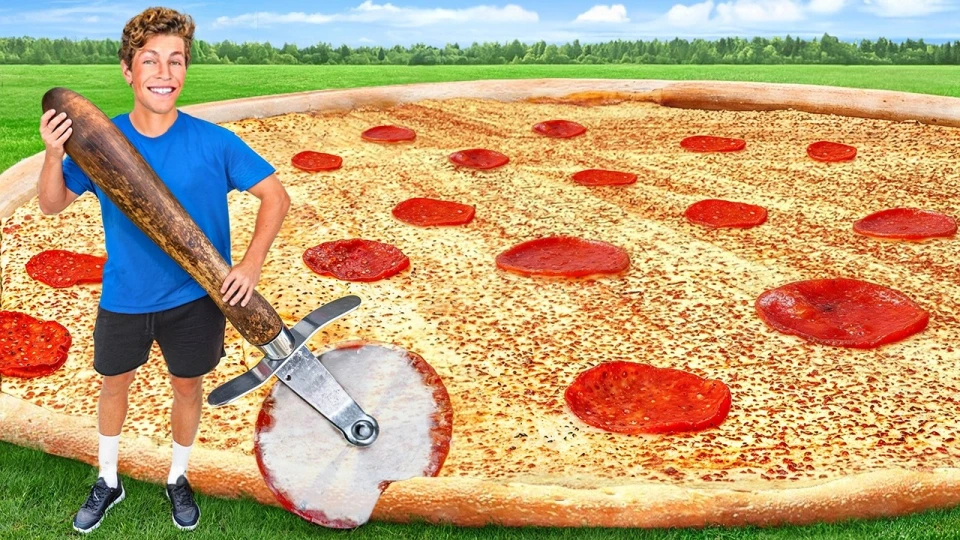Young man in blue shirt and black shorts holding an oversized wooden-handled pizza cutter next to a giant pepperoni pizza on grass under a blue sky.