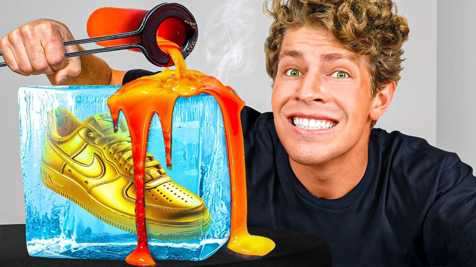 Man with curly hair and green eyes smiling widely while pouring orange molten material over a golden Nike shoe encased in a blue ice block on a black surface.