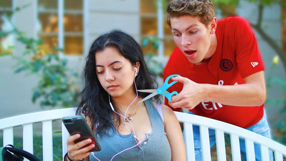 A young woman sitting on a white bench, looking at her phone with earphones plugged in, while a young man behind her is cutting the earphone wires with a pair of blue scissors, appearing surprised or excited.