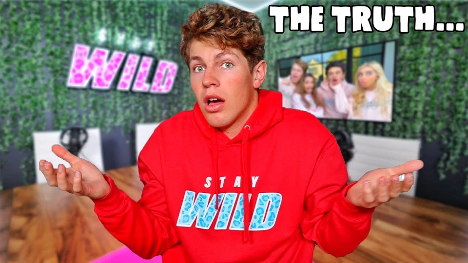 Young man in a red hoodie with 'STAY WILD' printed on it, shrugging with an expression of surprise or confusion in a modern room with green walls and a TV showing four people, with 'THE TRUTH...' and 'WILD' text overlayed.
