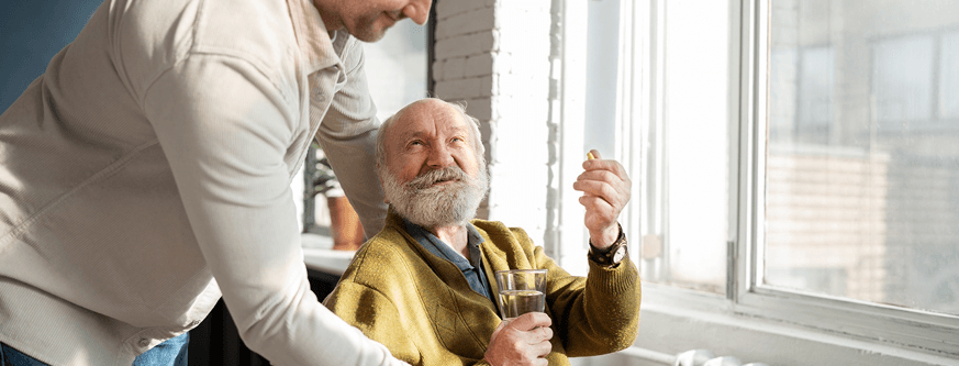 Elderly man sitting by a window holding a glass of water and a pill, smiling at a younger man assisting him.