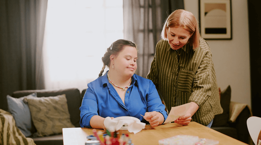 A woman in a green striped shirt showing a paper to a seated young woman with Down syndrome wearing a blue shirt and beaded necklace.