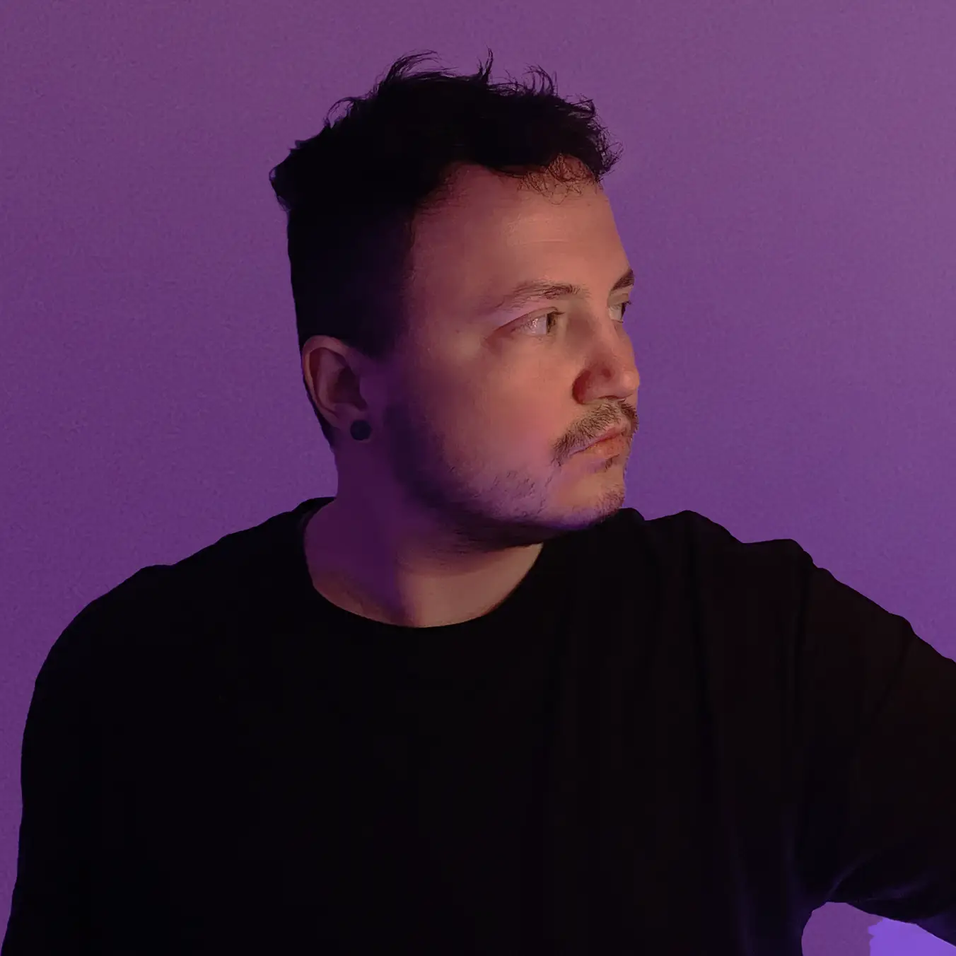Porträt eines Mannes mit kurzem dunklem Haar und Bart vor lila Hintergrund, der nach rechts schaut.