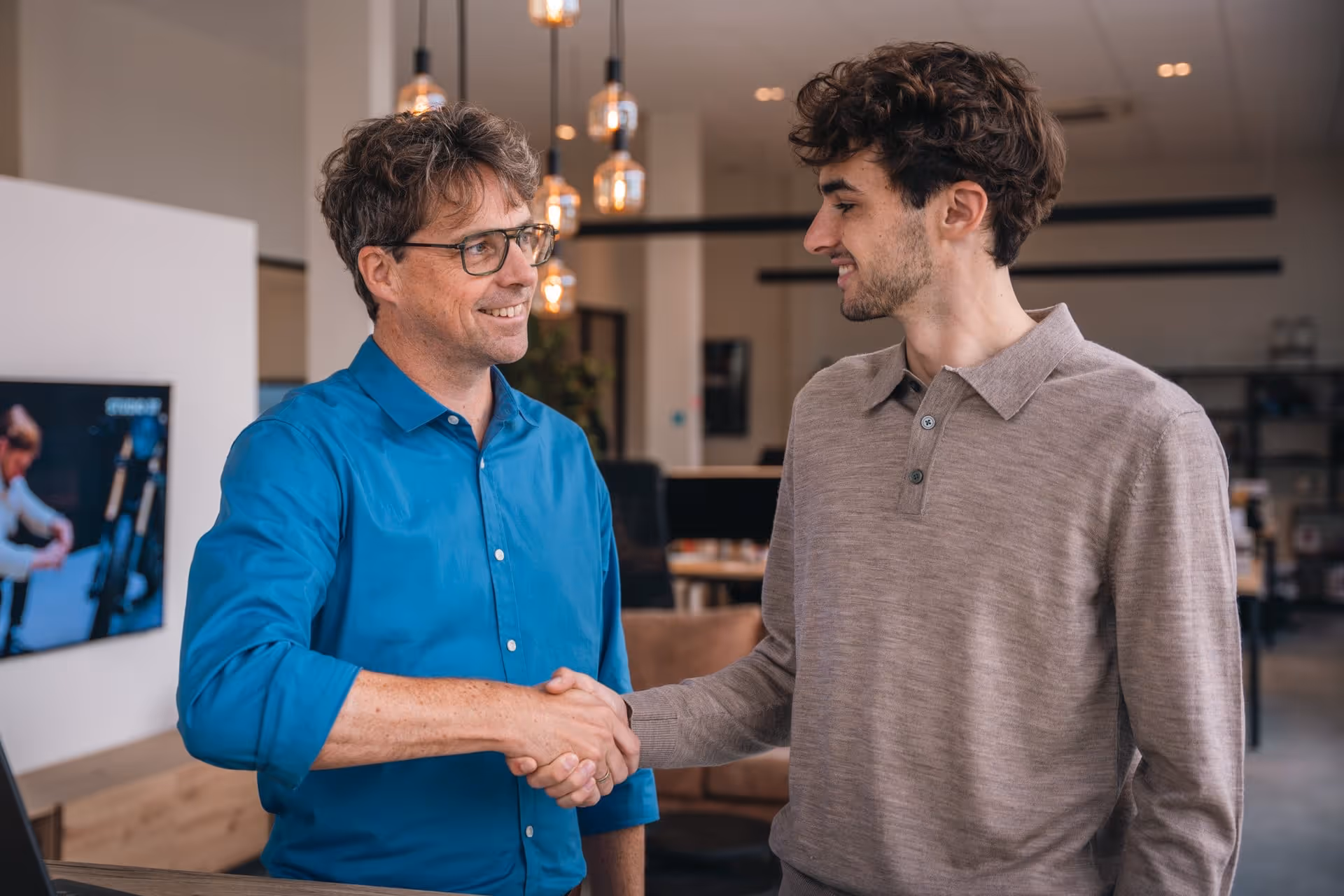 Tom draagt blauw hemd, bril; schudt de hand met een andere man; draagt bruine polo.