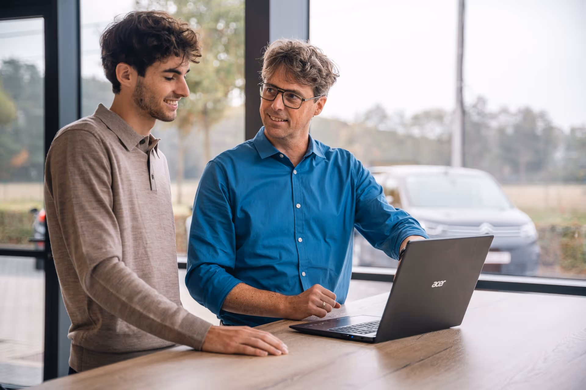 Tom toont iets op zijn laptop aan een andere man en hij wijst naar zijn scherm.