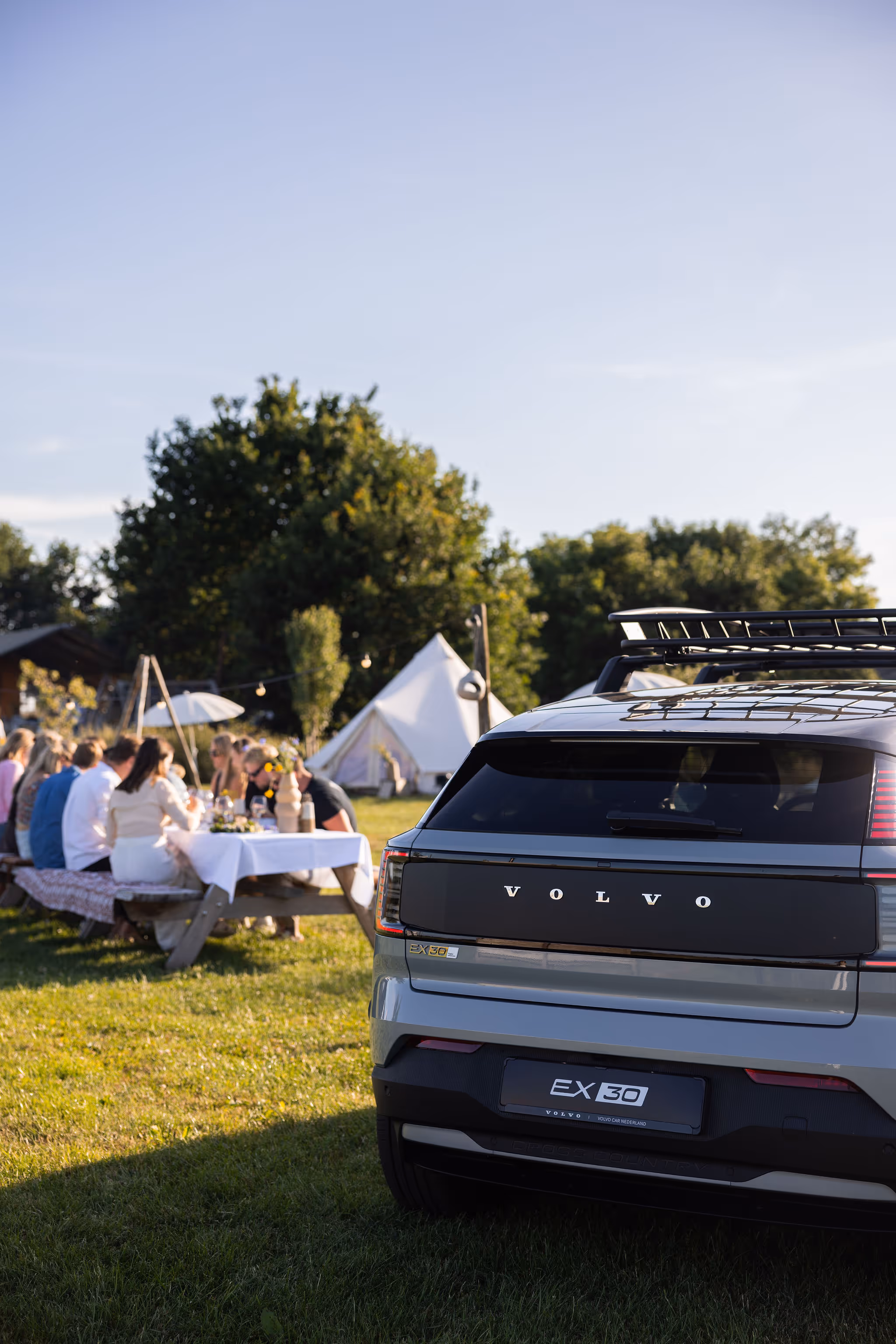 A Volvo EX90 SUV is parked on grass near a group of people sitting at outdoor tables, with a tent and trees visible in the background on a sunny day.