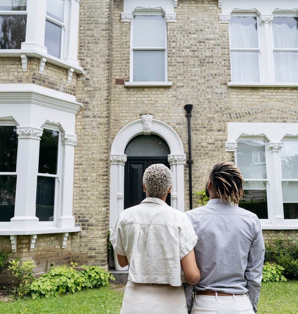 A couple stands together in front of a house, smiling and holding hands, with a clear blue sky in the background.