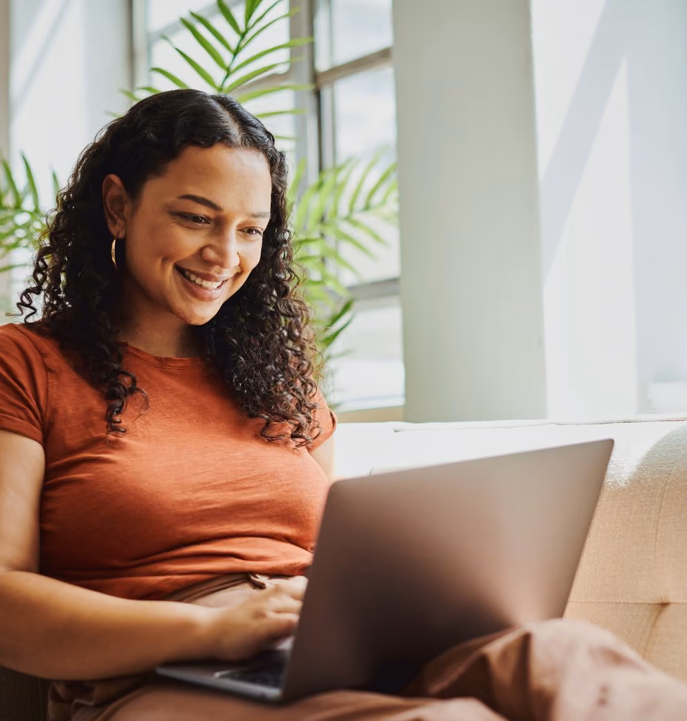A woman sitting on a couch, focused on her laptop, with a cozy living room setting in the background.