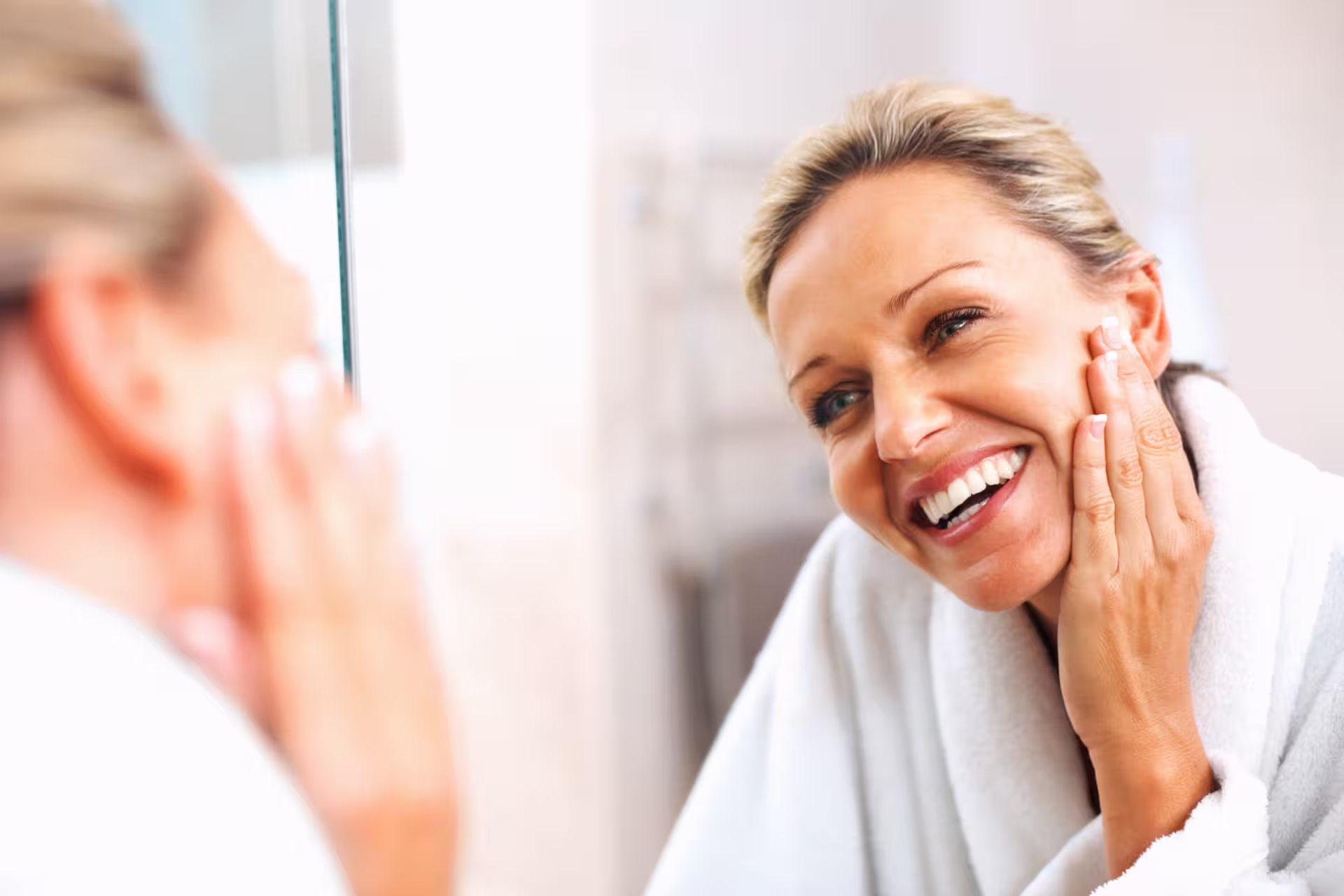 A woman in a bathrobe examines her reflection in the mirror, focusing on her face.