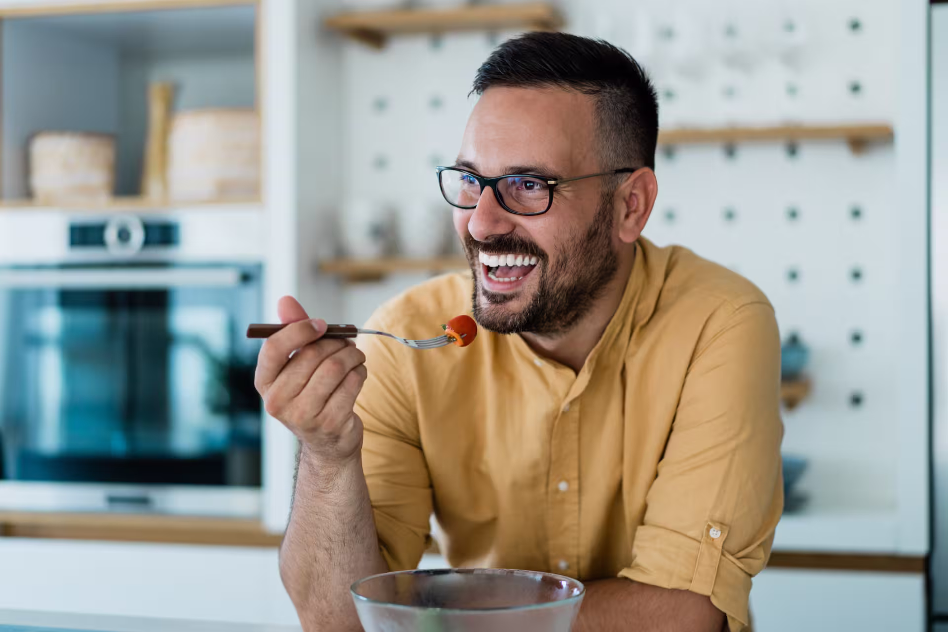 A man with glasses enjoys a fresh fruit while seated in a modern kitchen.