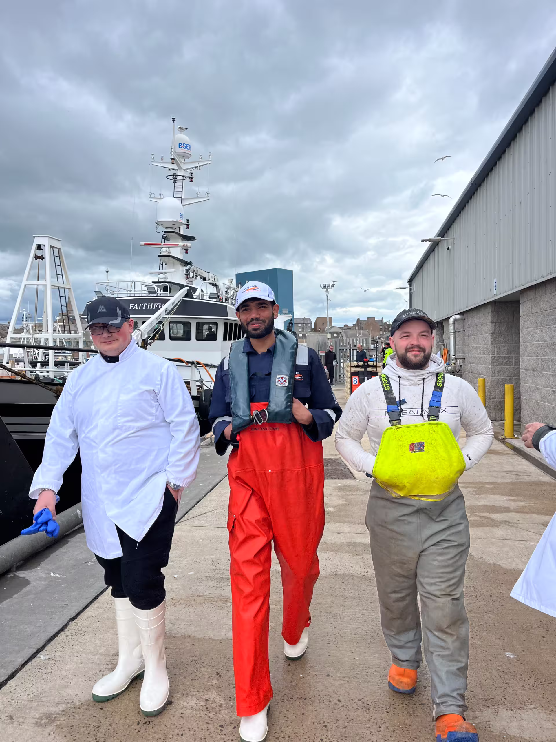 Three individuals wearing orange pants and white shirts stand together, smiling in a casual setting.
