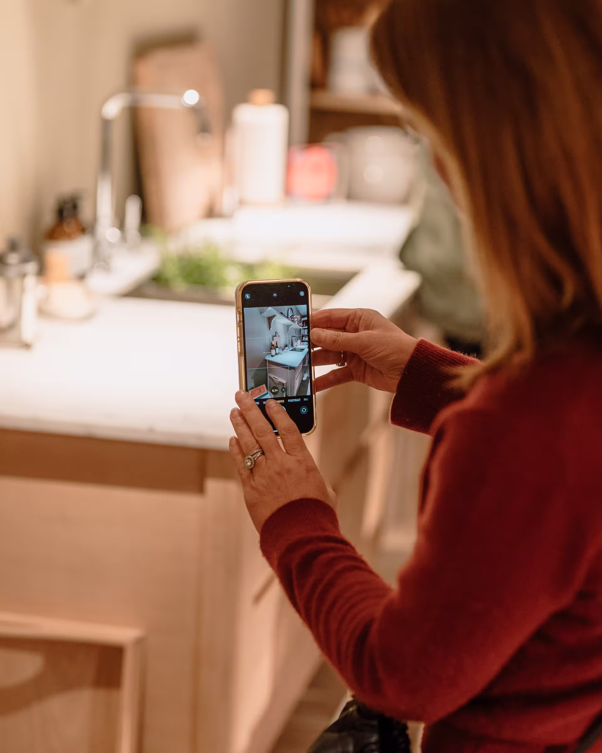 A woman taking a selfie in her kitchen, smiling while holding her phone in front of her.