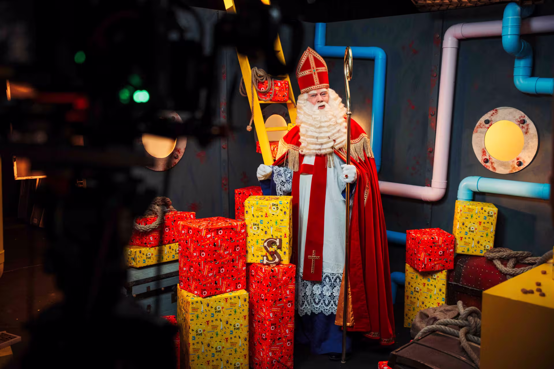 A man dressed as Santa Claus poses cheerfully in front of a camera, showcasing his festive red suit and white beard.