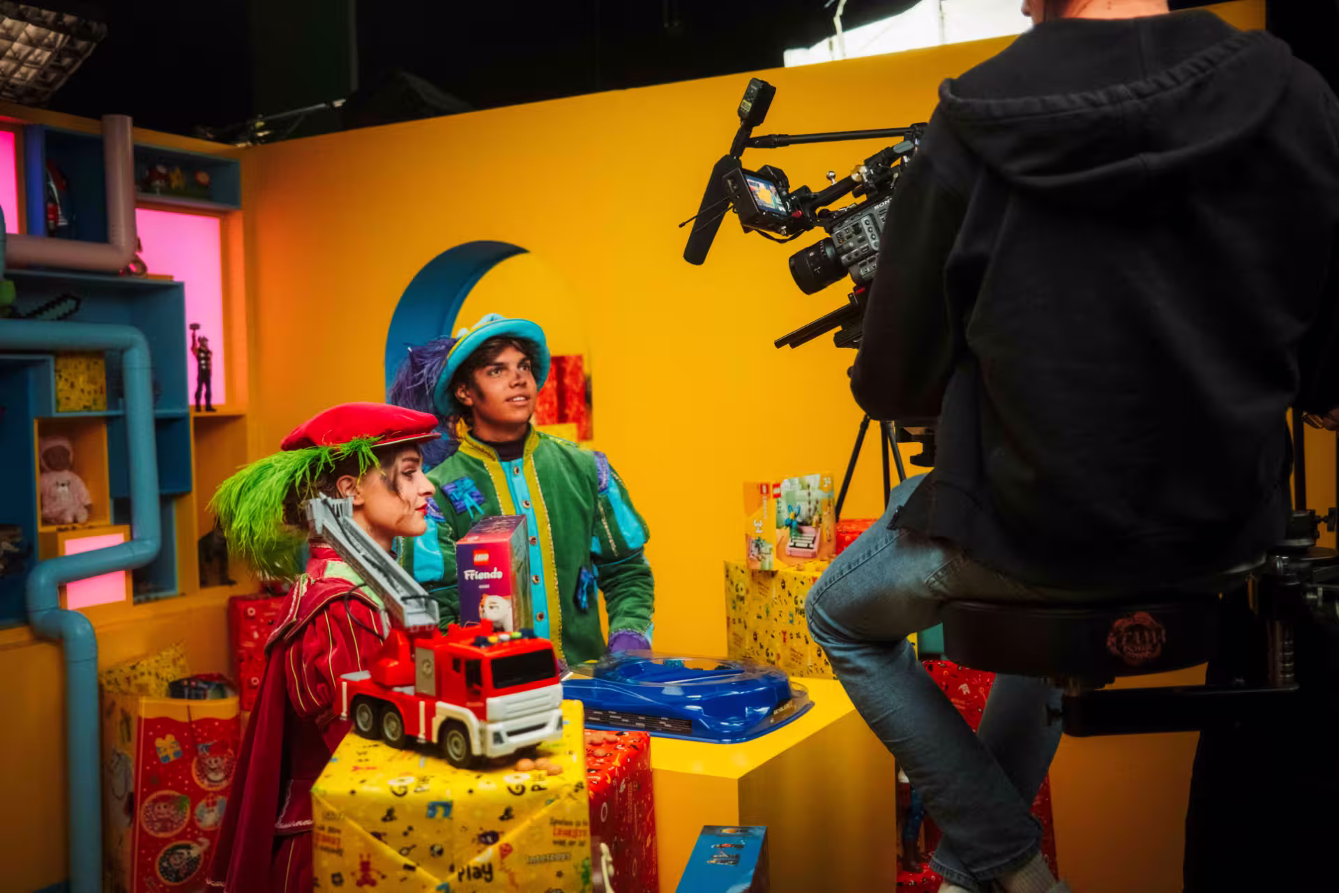 A man and woman in colorful clown costumes are filming a video together, smiling and posing for the camera.