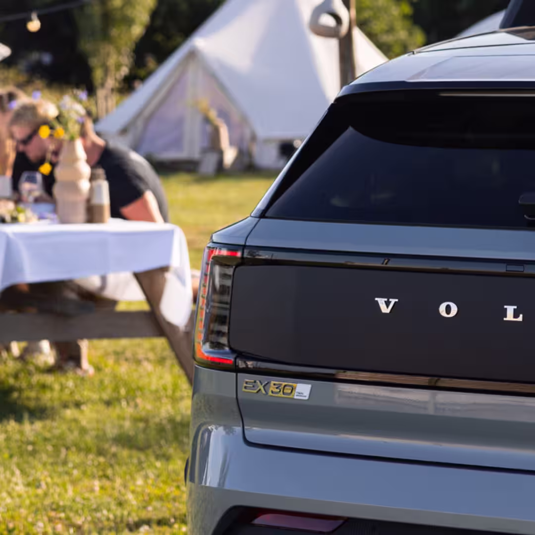 A Volvo EX90 SUV is parked on grass near a group of people sitting at outdoor tables, with a tent and trees visible in the background on a sunny day.