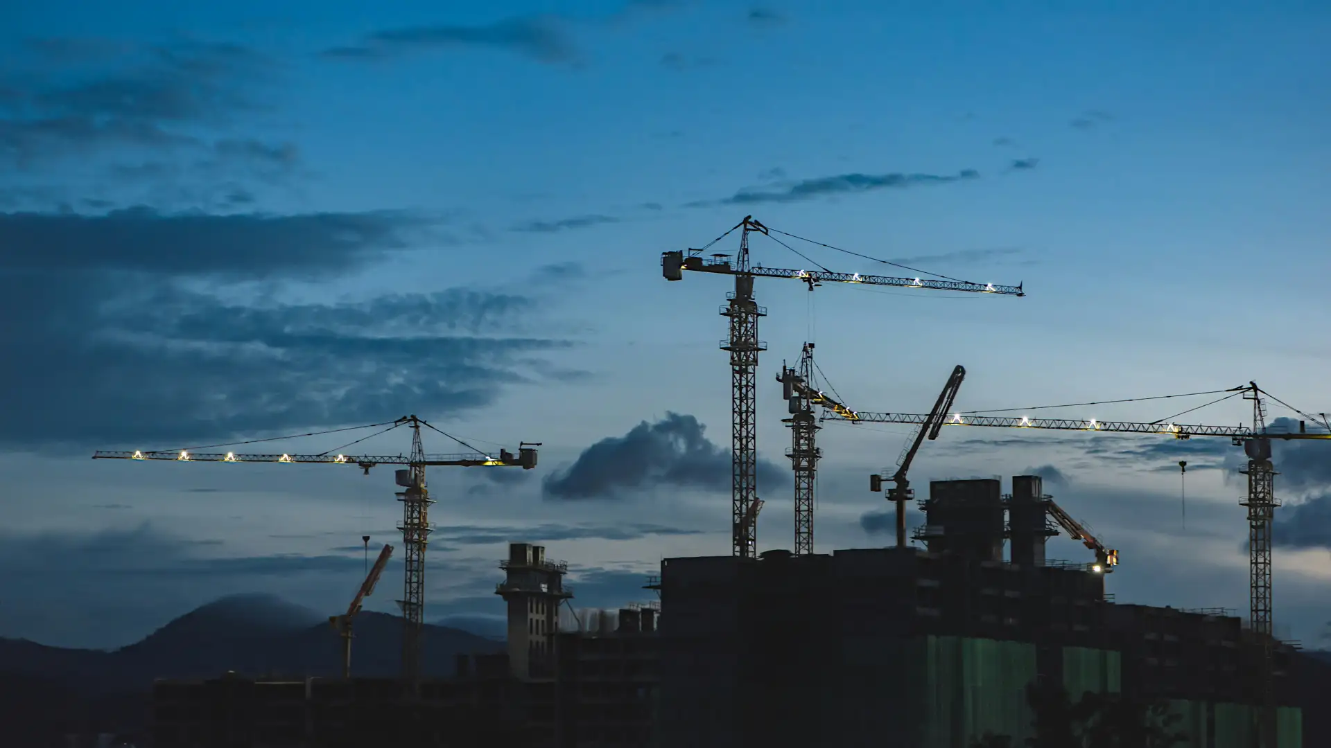 A silhouette of several large construction cranes against a deep blue twilight sky, with a dark building under construction in the foreground and distant mountains.