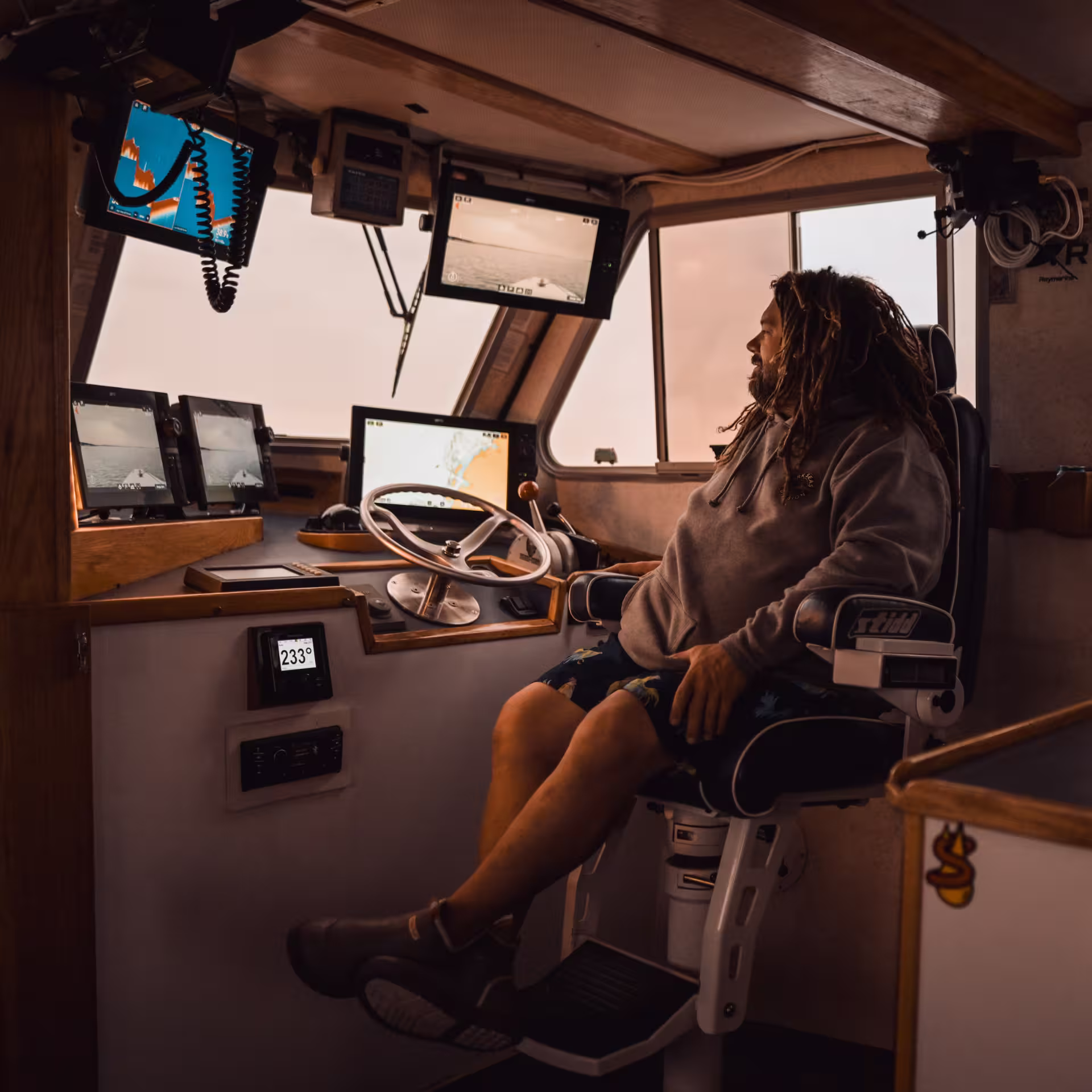 A man with long dreadlocks sitting in the captain's chair of a fishing boat wheelhouse, surrounded by multiple Raymarine chartplotter and sonar screens, with the cabin walls covered in fishing and outdoor brand stickers.