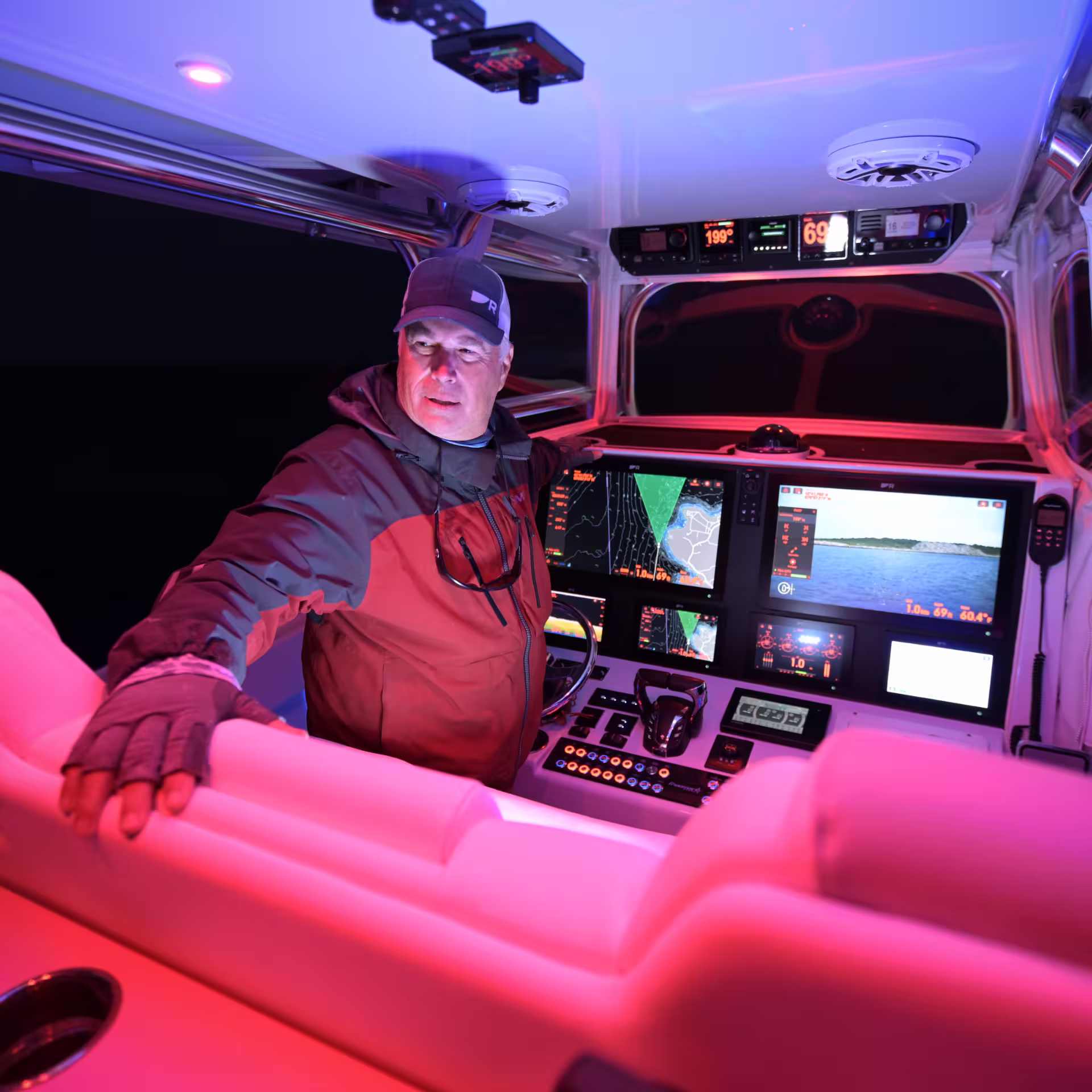 A boat captain in a dark jacket and Raymarine cap sitting at a helm station at night, illuminated by pink and blue ambient lighting, with multiple Raymarine multifunction displays showing charts, sonar, and navigation data