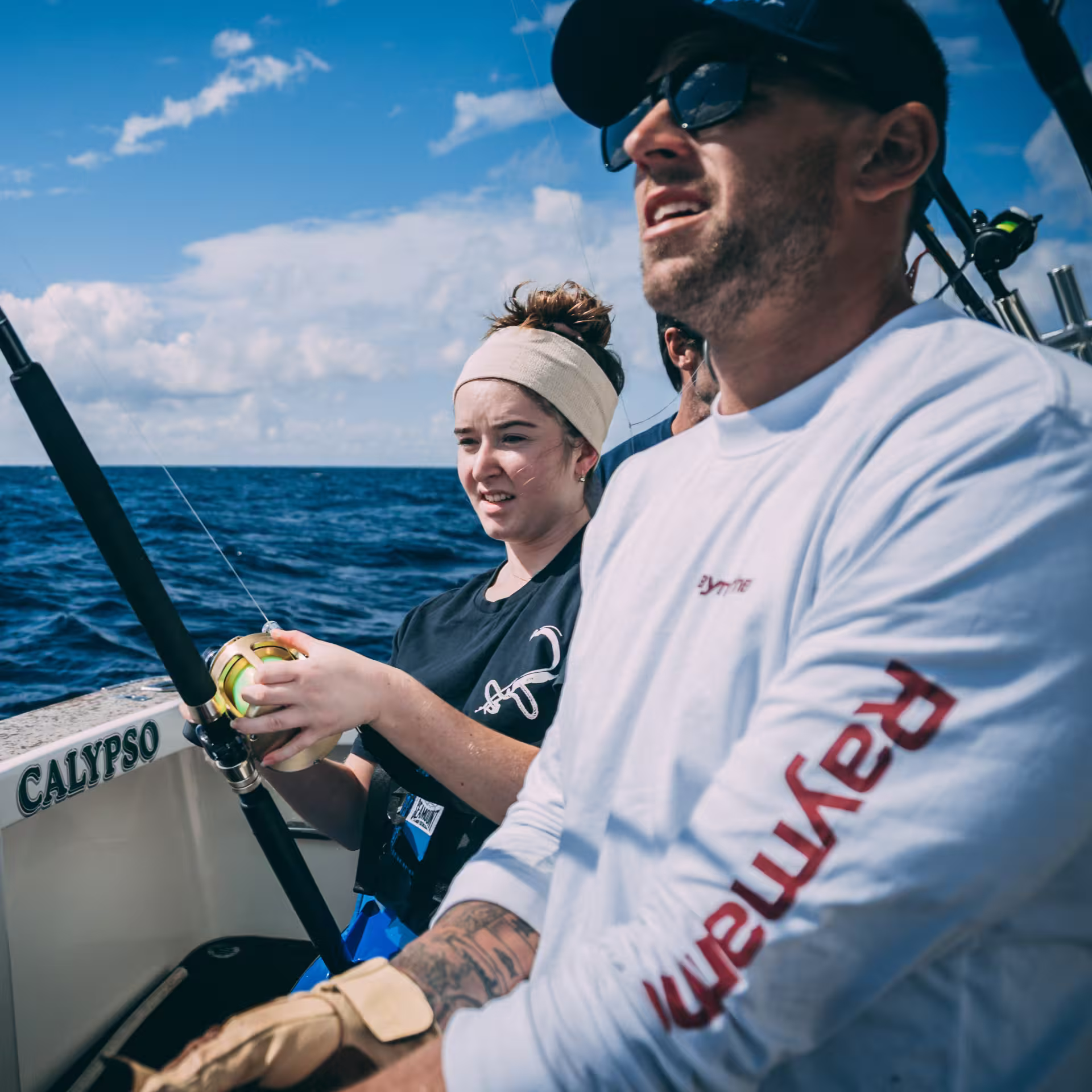 A young woman and a man fishing together on the stern of a boat named "Calypso" on a deep blue ocean under a partly cloudy sky. The man wears a white long-sleeve Raymarine shirt and sunglasses; the woman holds a fishing rod and reel