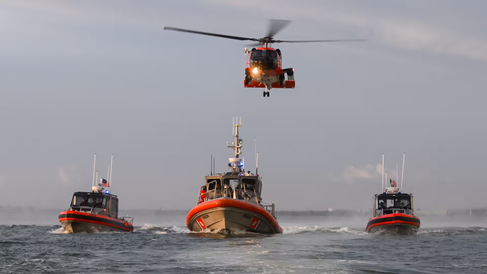 Three U.S. Coast Guard response boats in orange-and-black livery speeding across choppy water in formation, with an orange Coast Guard helicopter hovering directly above the center vessel, its searchlight illuminated