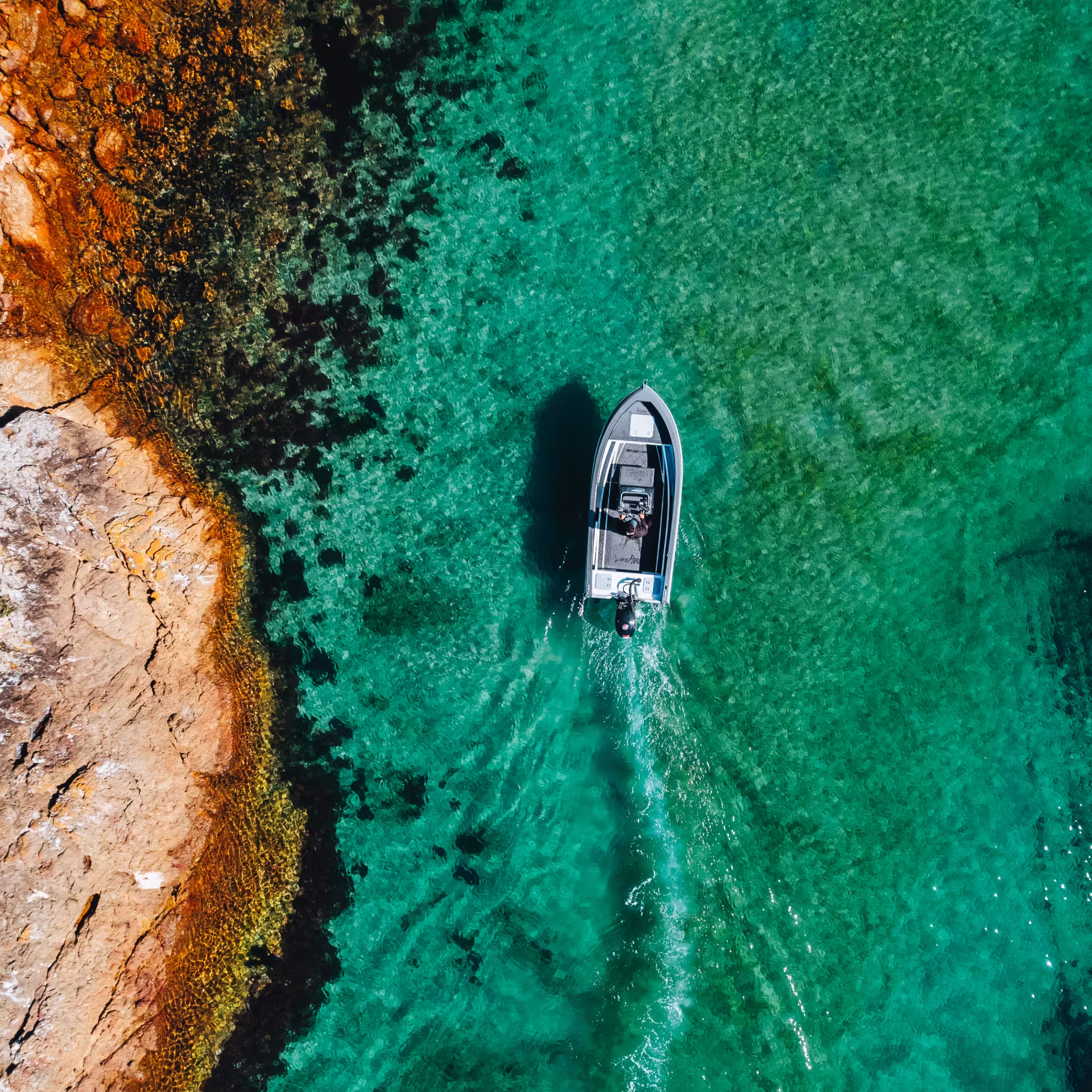 Top-down aerial view of a small aluminium boat motoring slowly through crystal-clear turquoise water near an orange-brown rocky shoreline