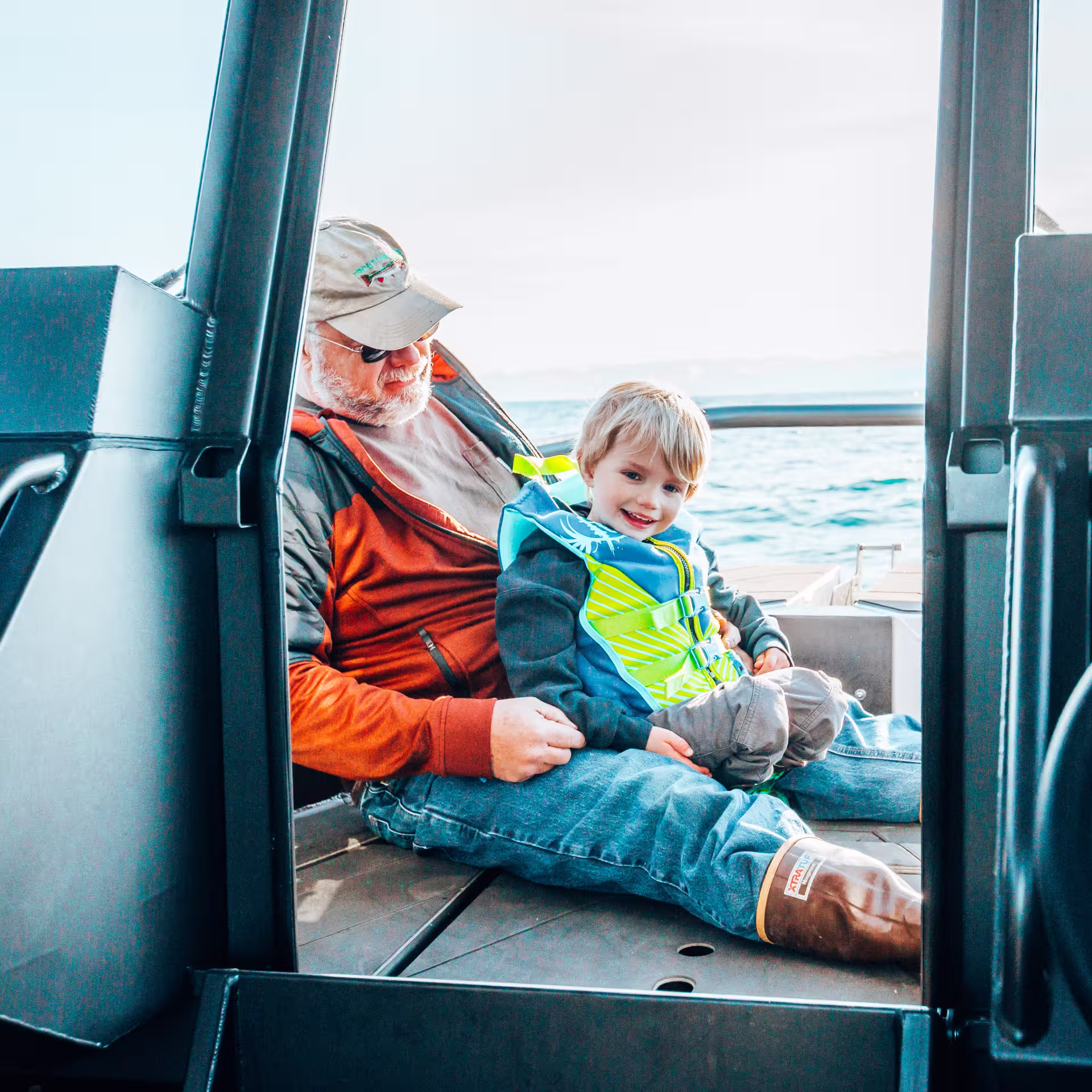 Grandfather and young child wearing a life jacket sitting together on a boat, with Raymarine navigation displays and a steering wheel visible in the foreground