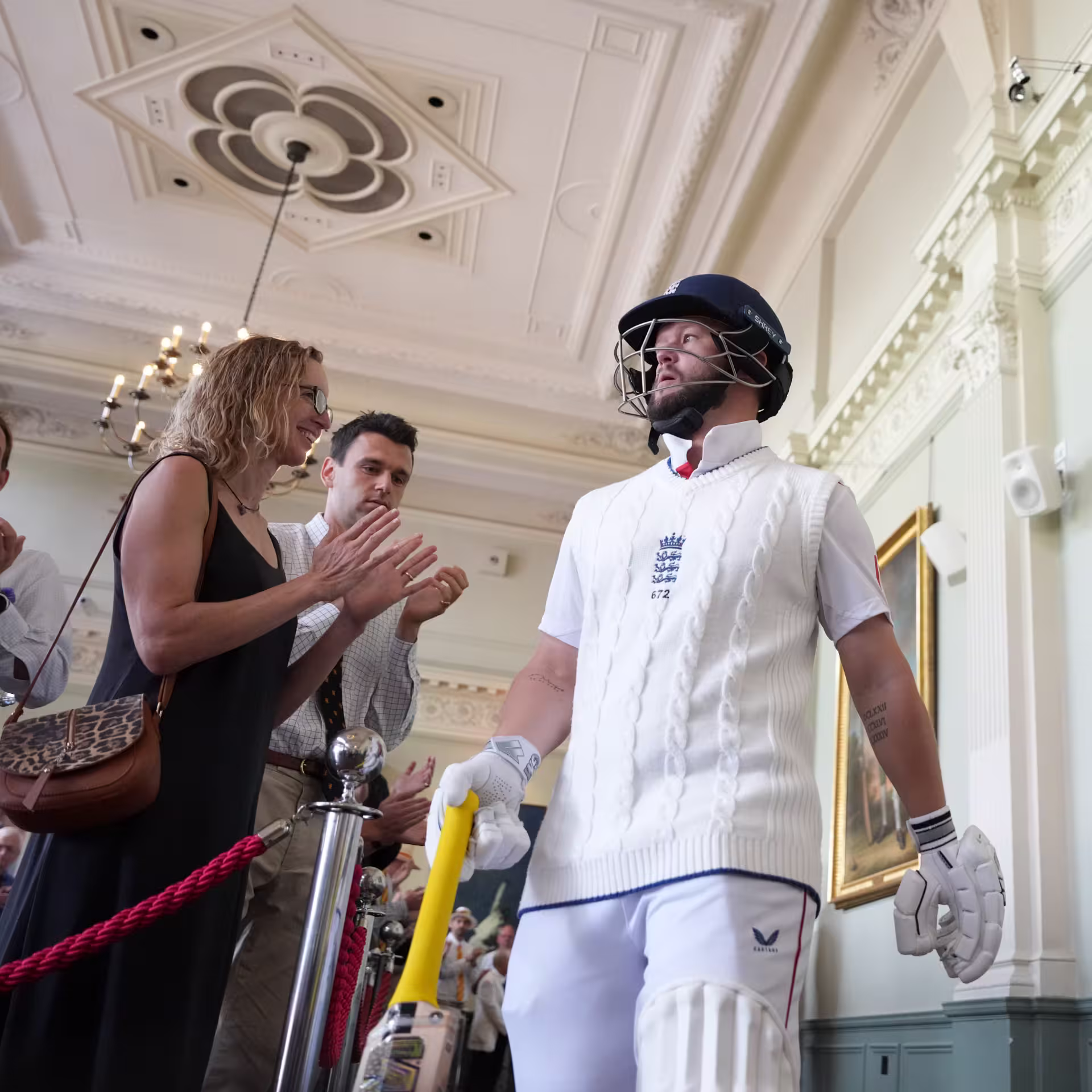 England batsman in whites and helmet walking through the Long Room at Lord's, applauded by spectators, with cricket paintings on the walls